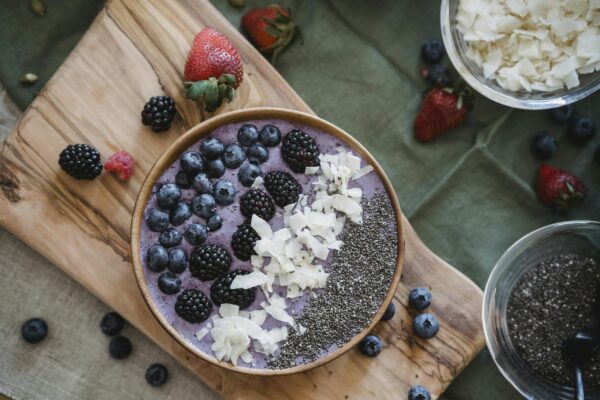 A healthy berry smoothie bowl topped with fresh fruits, seeds, and coconut shavings on a wooden board.