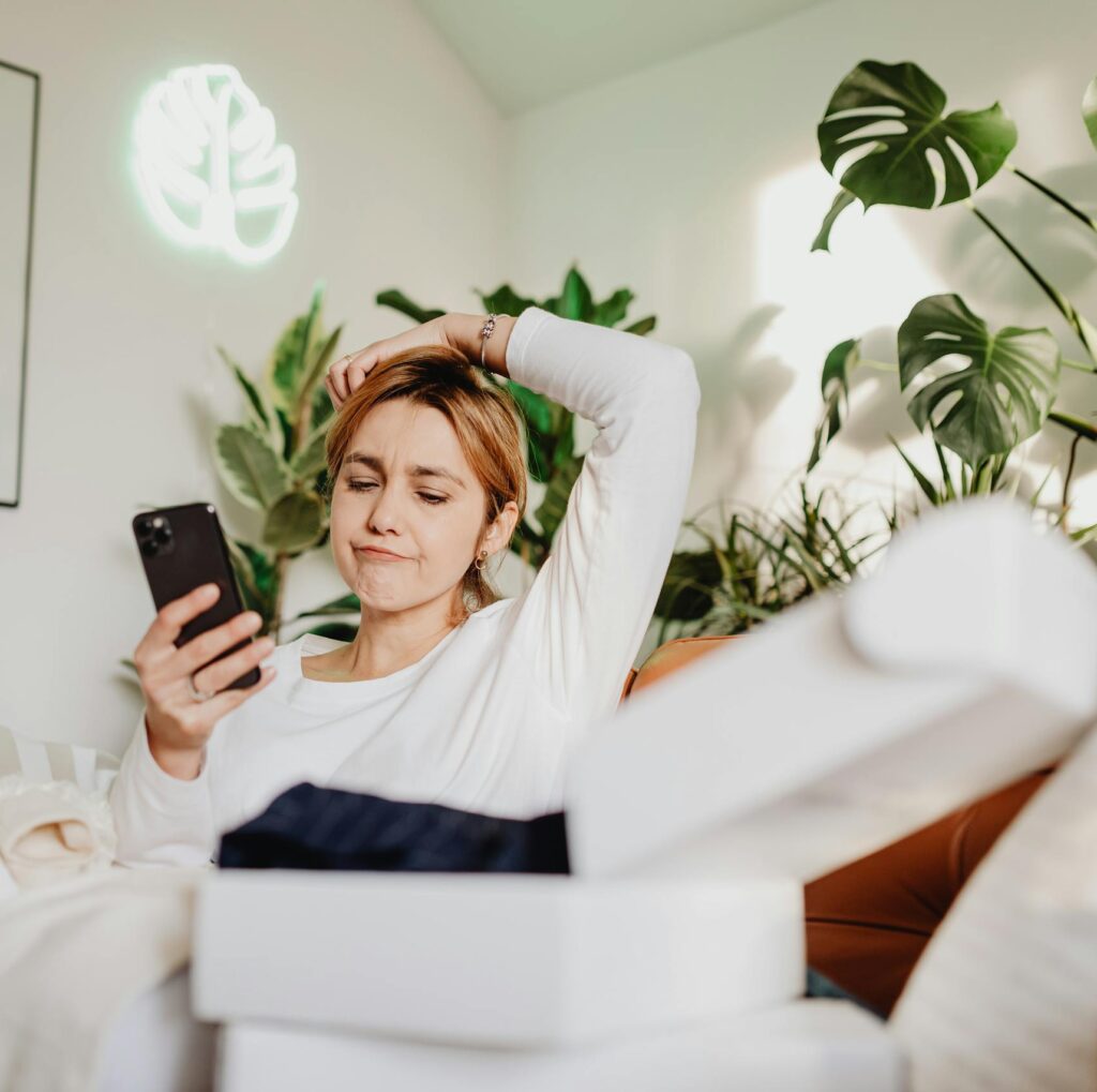 A woman sitting on a couch surrounded by plants, using her phone with a thoughtful expression.