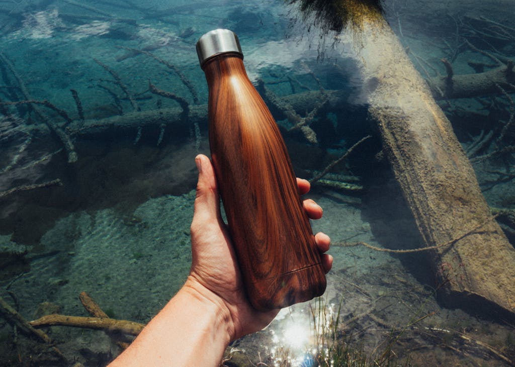 Close-up of a hand holding a wooden water bottle over a clear stream, showcasing eco-friendly lifestyle.