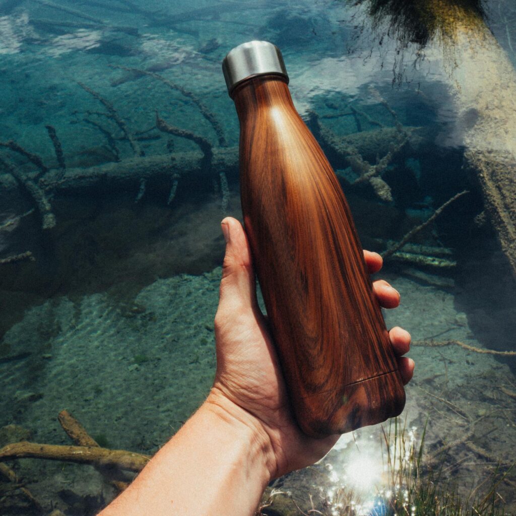 Close-up of a hand holding a wooden water bottle over a clear stream, showcasing eco-friendly lifestyle.