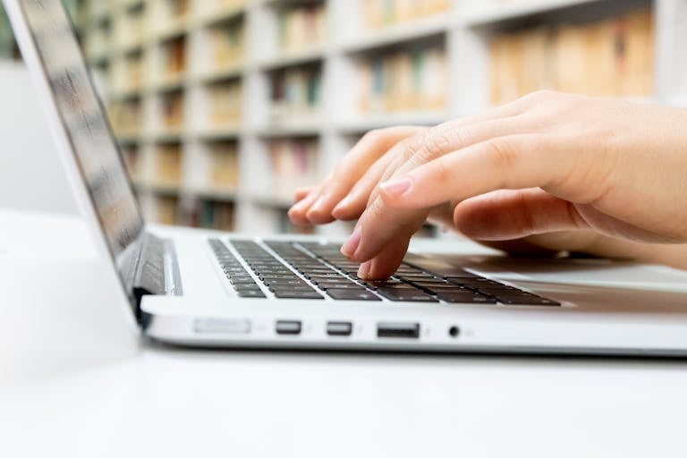 Close-up of hands typing on a laptop keyboard in a library setting.