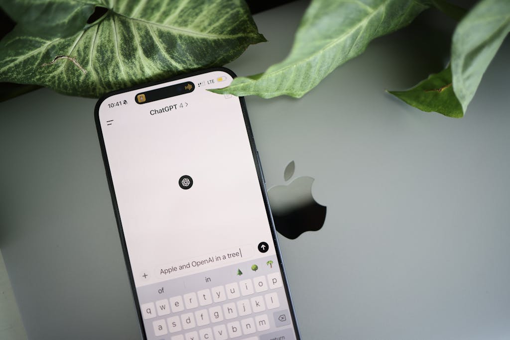 A smartphone shows a ChatGPT interface placed on an Apple laptop in a leafy environment.