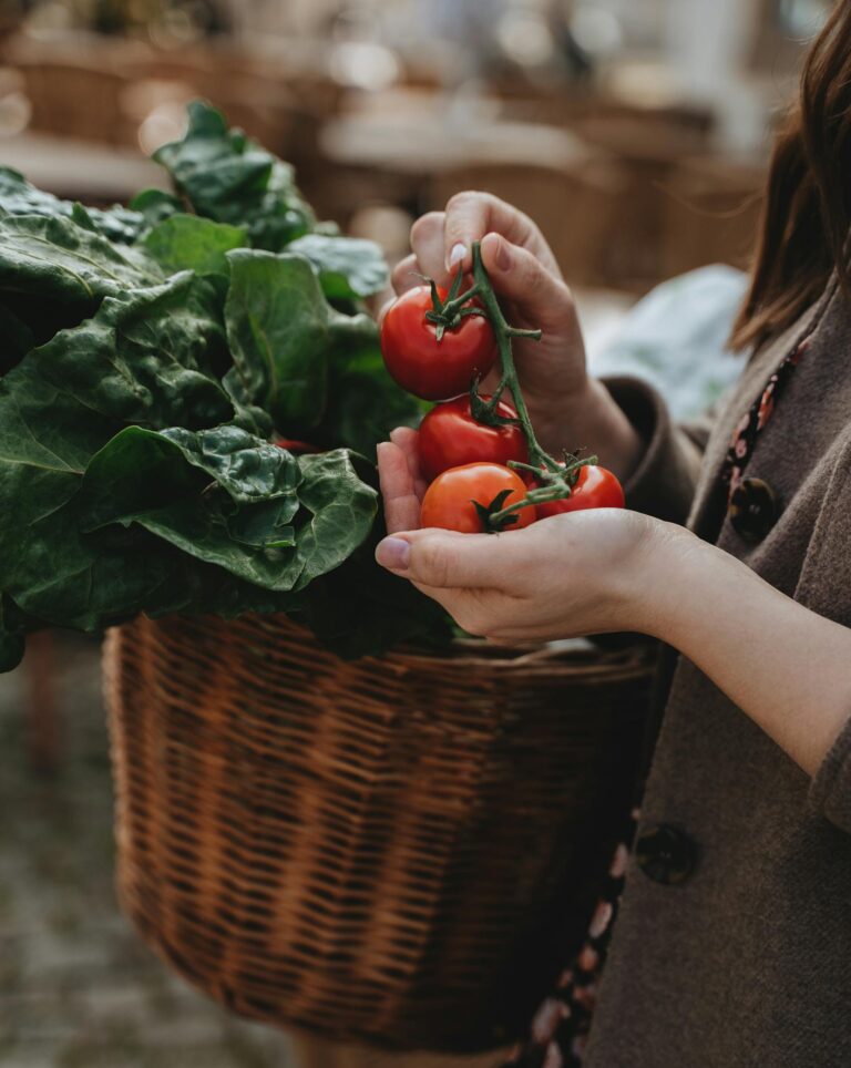 A woman holds ripe tomatoes with leafy greens in a wicker basket at a market.