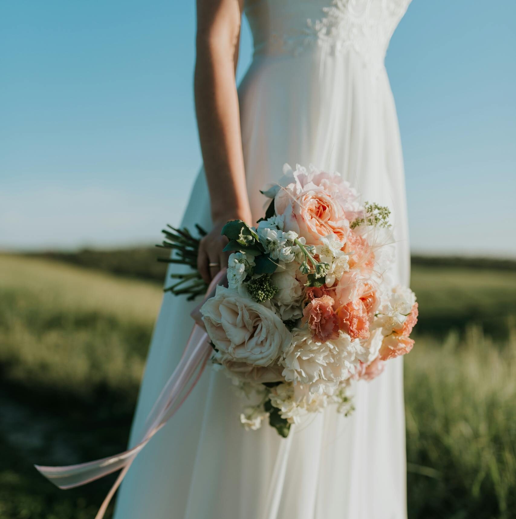 A bride in a flowing white gown holds a colorful bouquet in a sunny field.