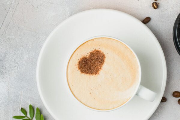 A cappuccino with a heart-shaped cocoa dusting on a saucer, surrounded by coffee beans.