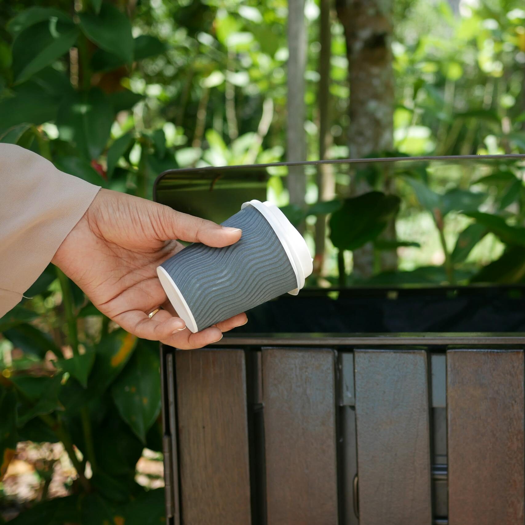 A person throws a biodegradable coffee cup into a recycling bin amidst greenery.