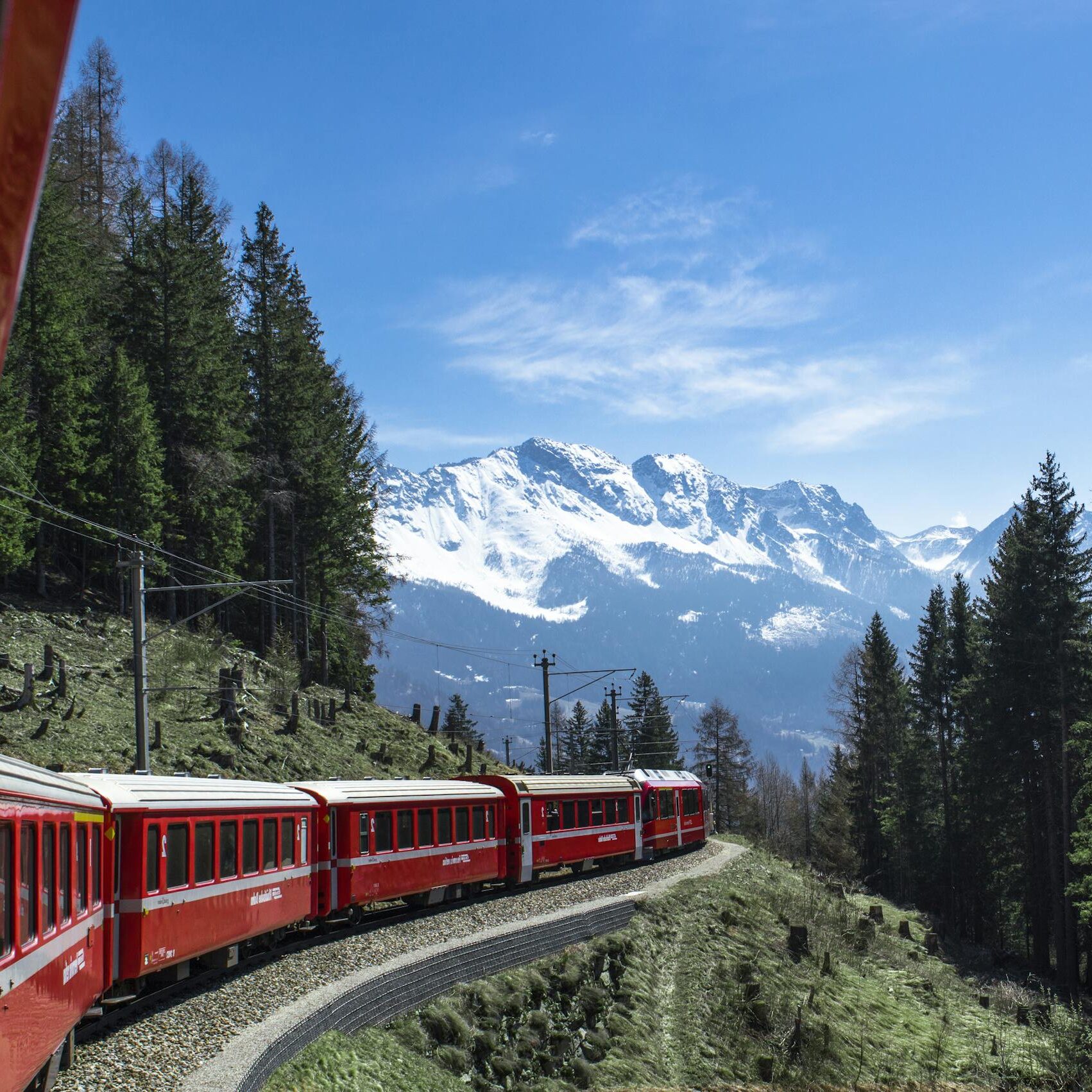 A red train travels through lush forests and snowy mountains under a clear blue sky in Switzerland.
