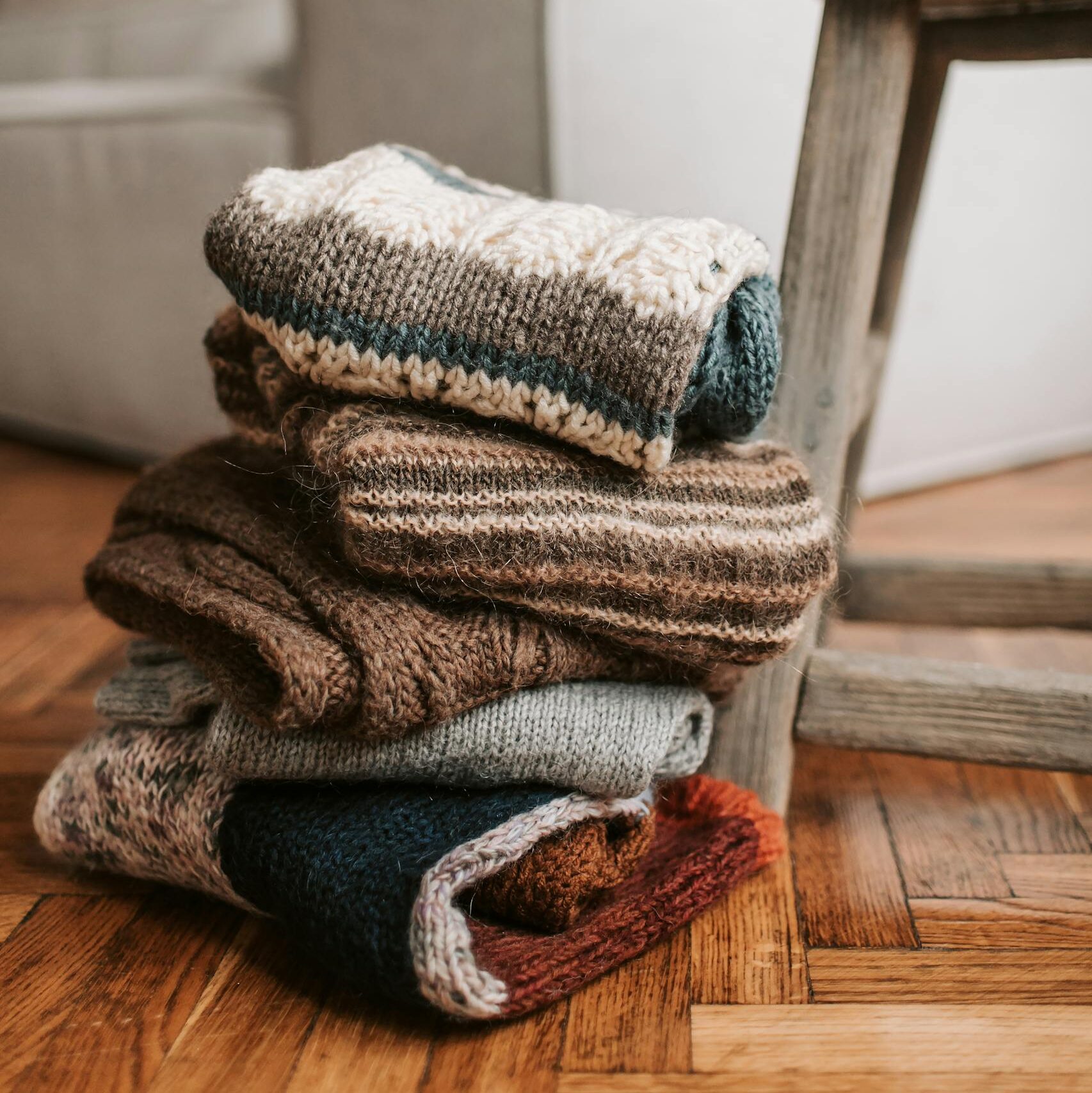 A stack of folded knitted scarves in earthy tones sitting on a rustic wooden chair indoors.
