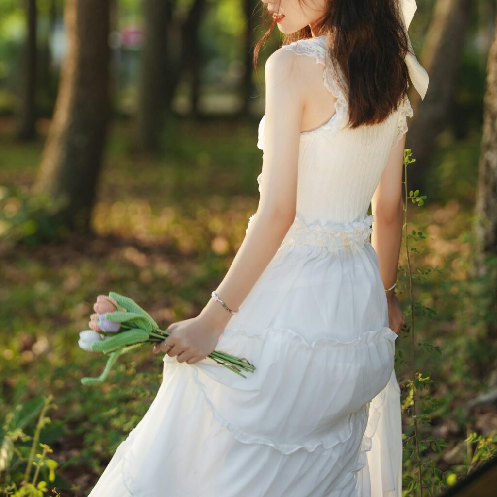 A woman in a white dress standing gracefully in a sunlit forest holding tulips.