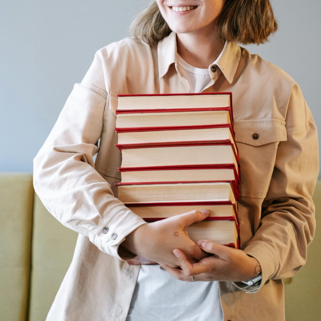 A young woman smiling while holding a stack of books indoors, embodying joy and education.