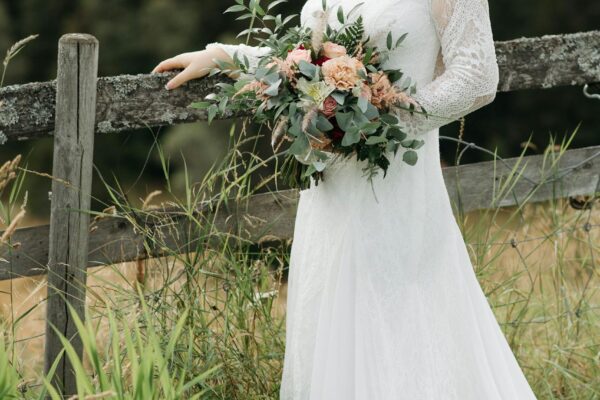 Bride in a white wedding dress holding a bouquet by a rustic fence with forest and mountains in the background.