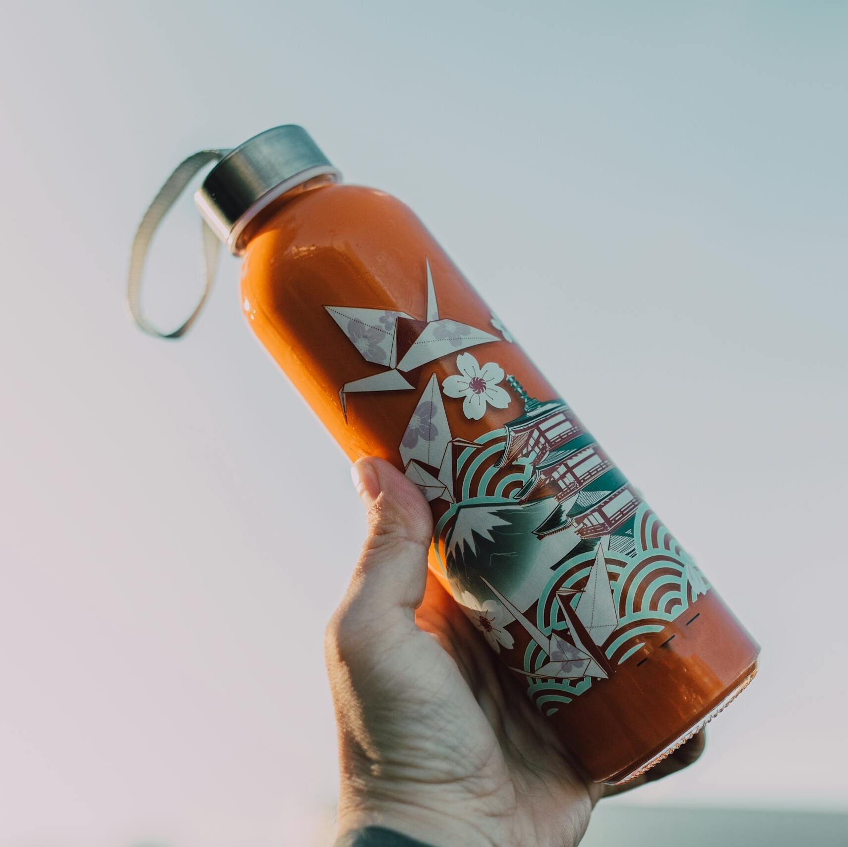 Close-up of a hand holding a decorative reusable water bottle against a clear sky.