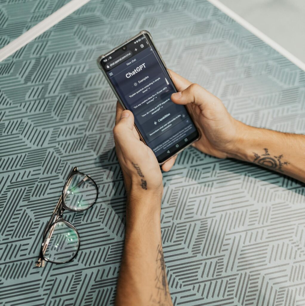 Hands holding smartphone displaying ChatGPT app near eyeglasses on table.