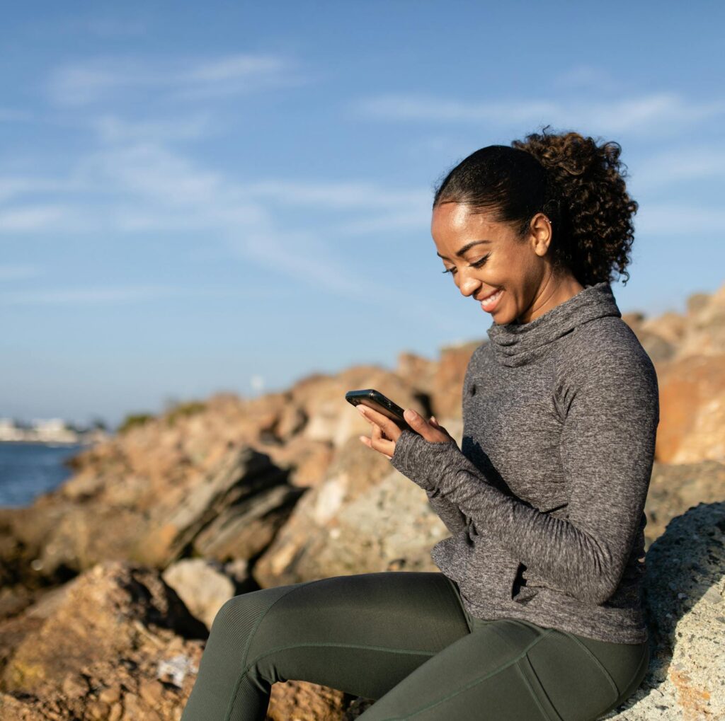 Smiling woman using smartphone on rocky shore, enjoying a sunny day.