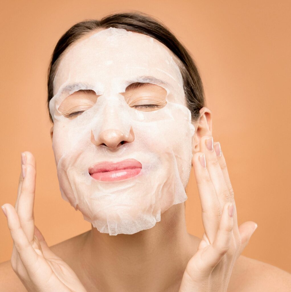 Woman smiling with a sheet face mask, embracing a skincare self-care moment.