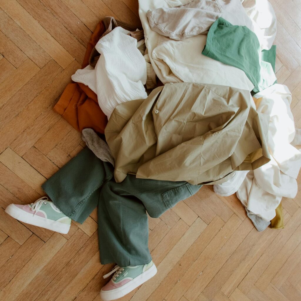 A child playfully hides under a pile of casual clothes on a parquet wooden floor.
