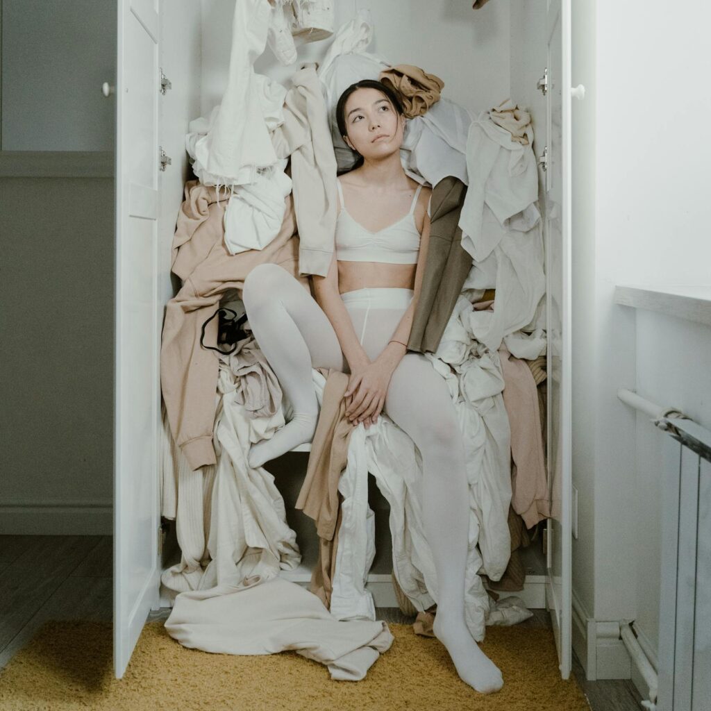 A contemplative woman sits surrounded by clothes in a messy wardrobe, evoking themes of consumerism and clutter.