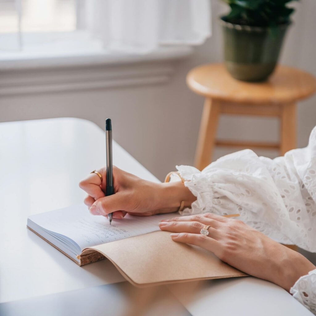Close-up of a woman writing in a notebook at a bright home office desk.