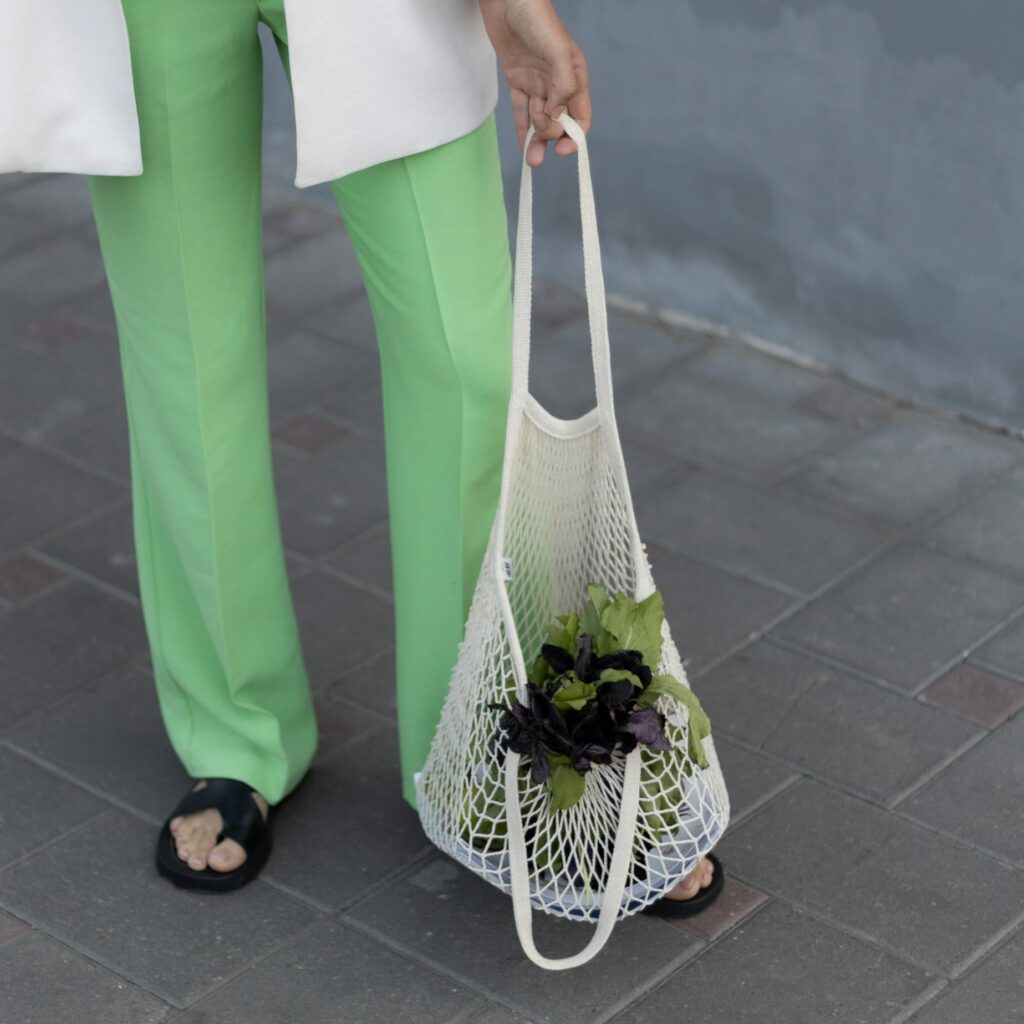 Fashionable individual holding a mesh bag with leafy greens on urban pavement.