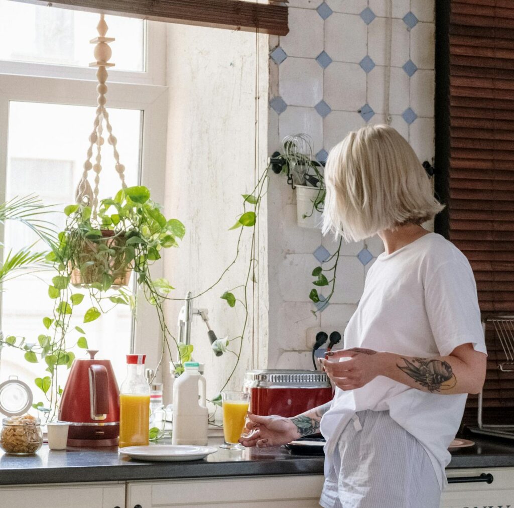 Woman with a tattoo preparing orange juice in a bright kitchen with indoor plants.