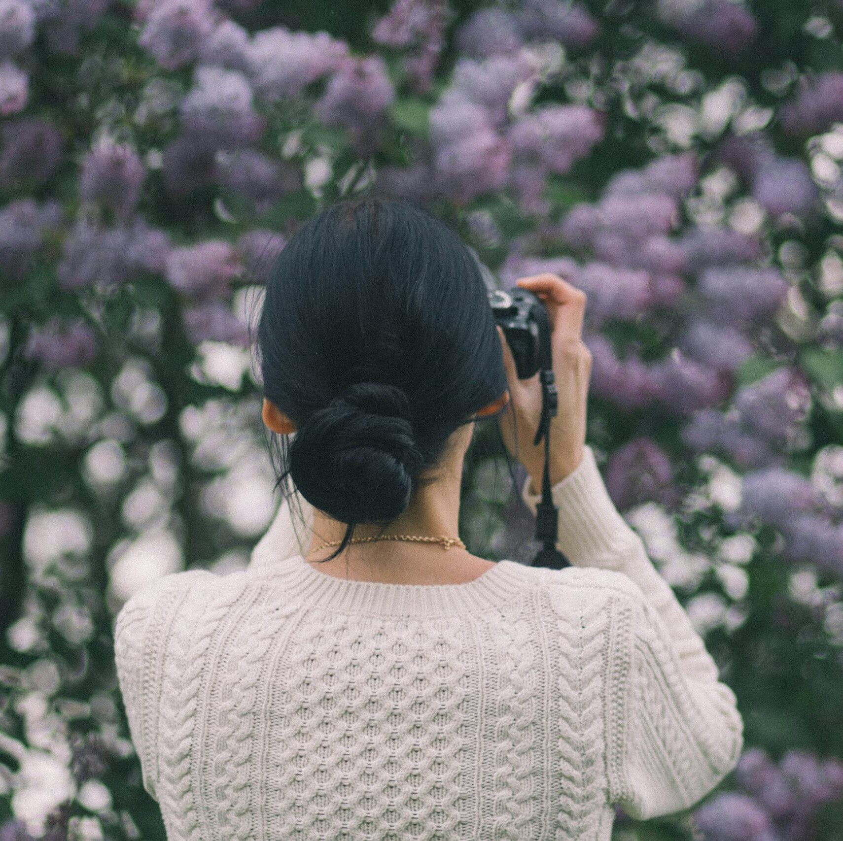 A woman with braided hair capturing vibrant lilac flowers in an outdoor garden during the day.