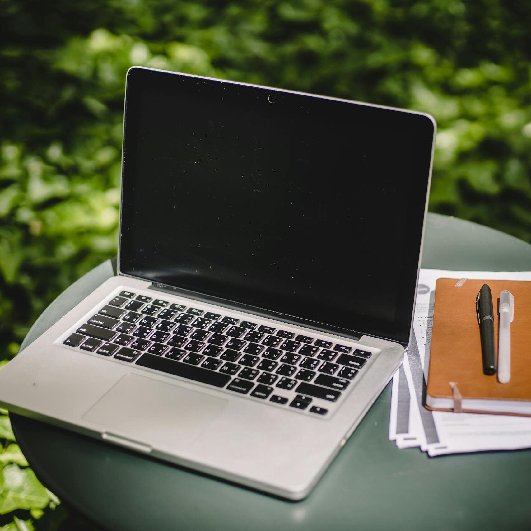 Laptop on a table outdoors with notebook and pen, ideal remote workspace.