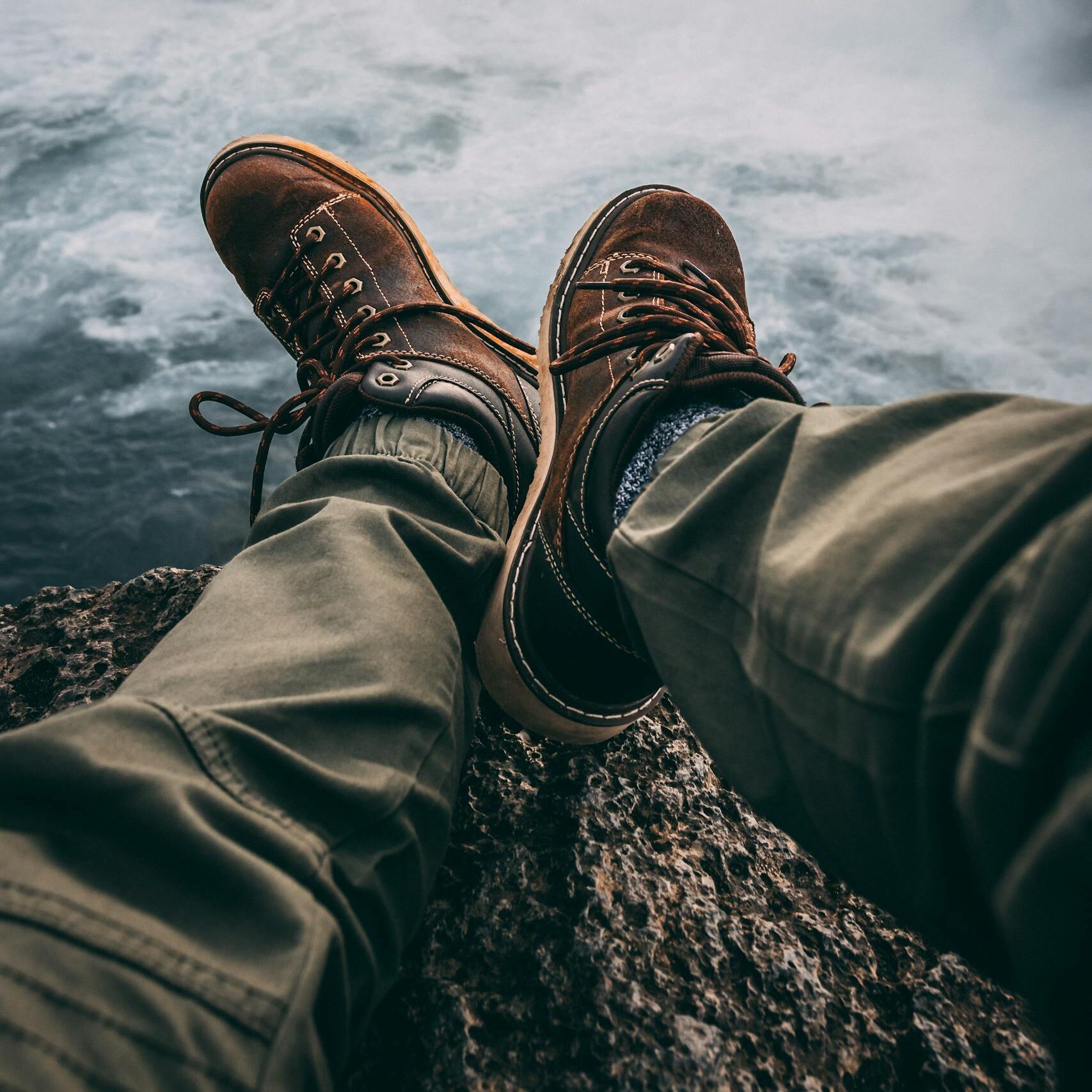 Person in hiking boots sitting at cliff's edge, overlooking breathtaking waterfall.
