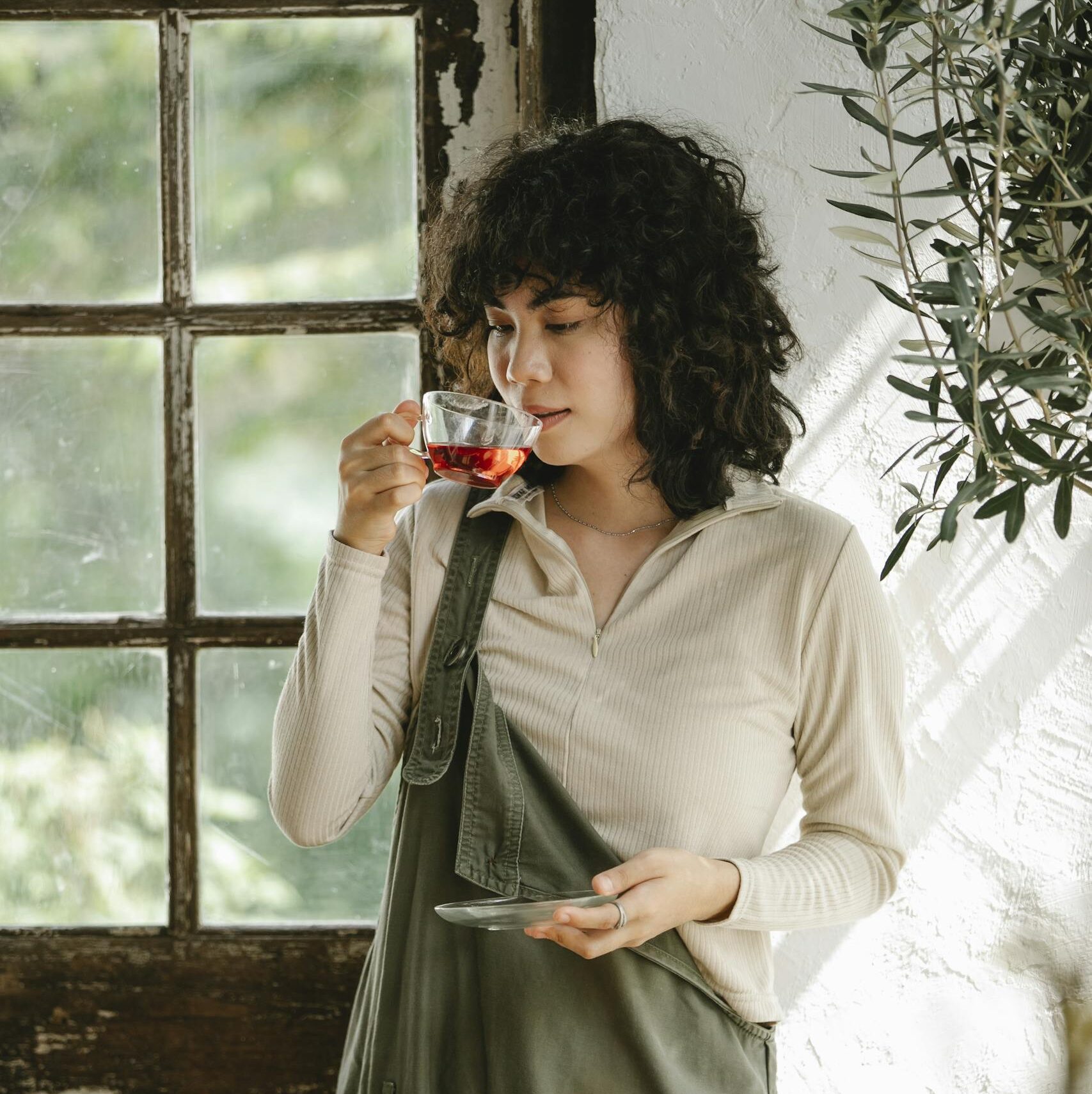 Serene ethnic woman drinking hot tea from glass cup standing at doorway of shabby rustic house