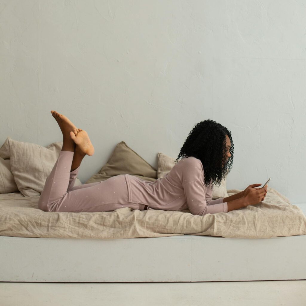 Woman in comfortable pajamas lying on bed, using phone in a minimalist bedroom setting.