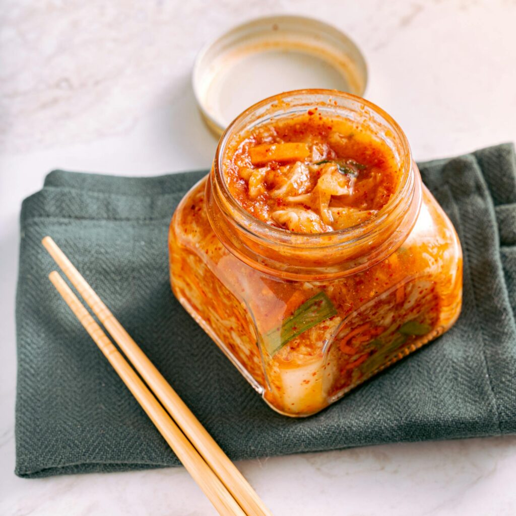A jar of traditional homemade kimchi on a marble surface with wooden chopsticks beside it.