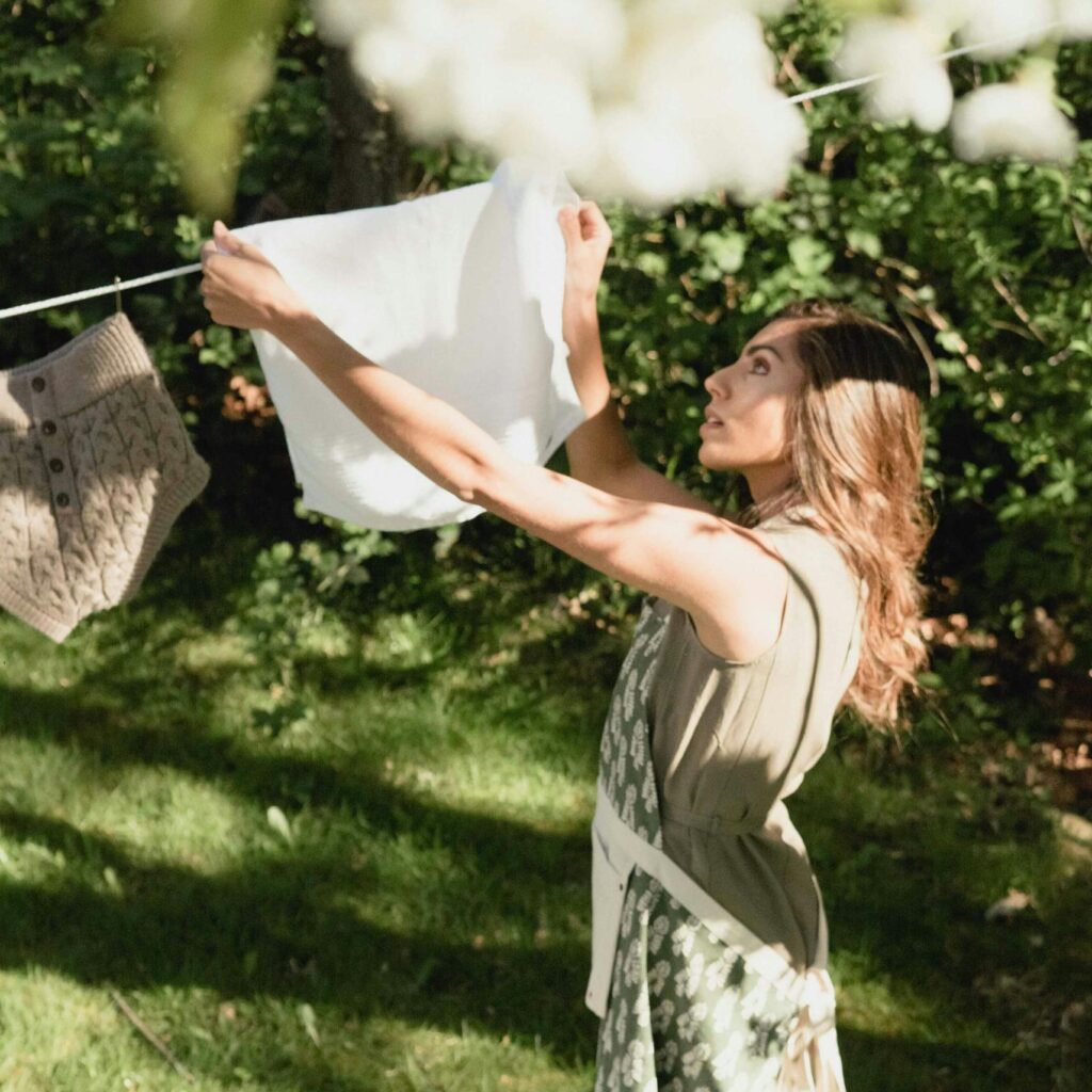 A woman hangs clothes on a line in a sunlit garden, enjoying a summer day.