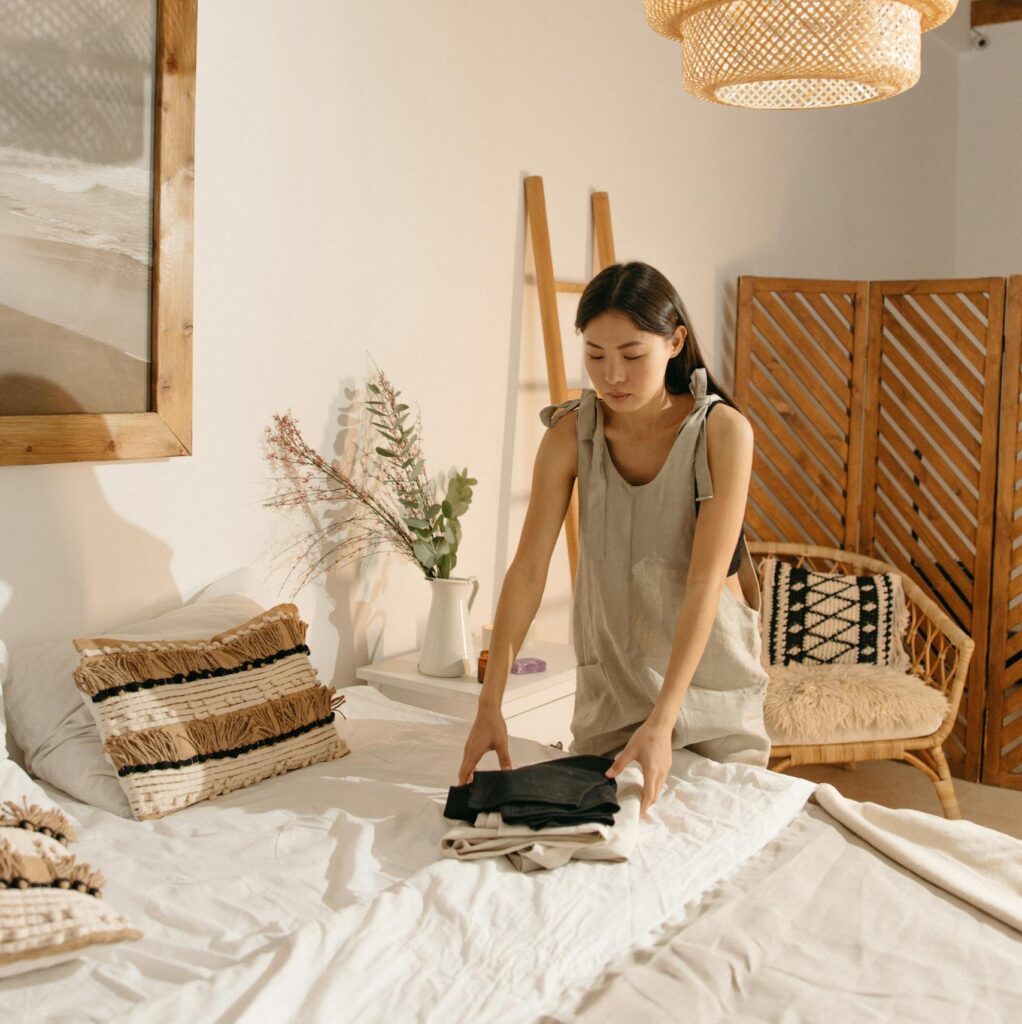 Asian woman folding clothes in a stylish, minimalist bedroom with warm décor and pendant lighting.