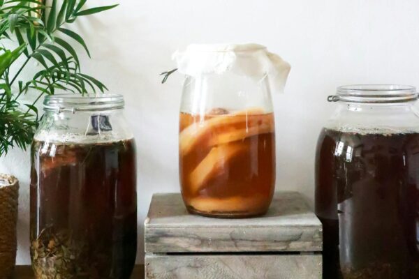 Glass jars with infused liqueurs with ingredients placed on wooden table near green plant near wall with decorations at home