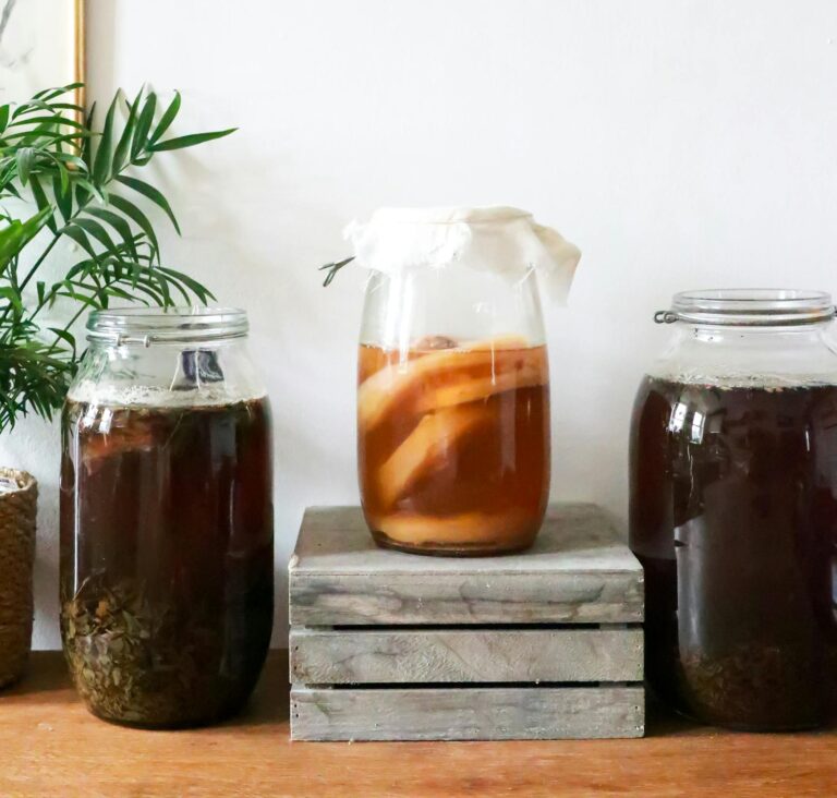 Glass jars with infused liqueurs with ingredients placed on wooden table near green plant near wall with decorations at home