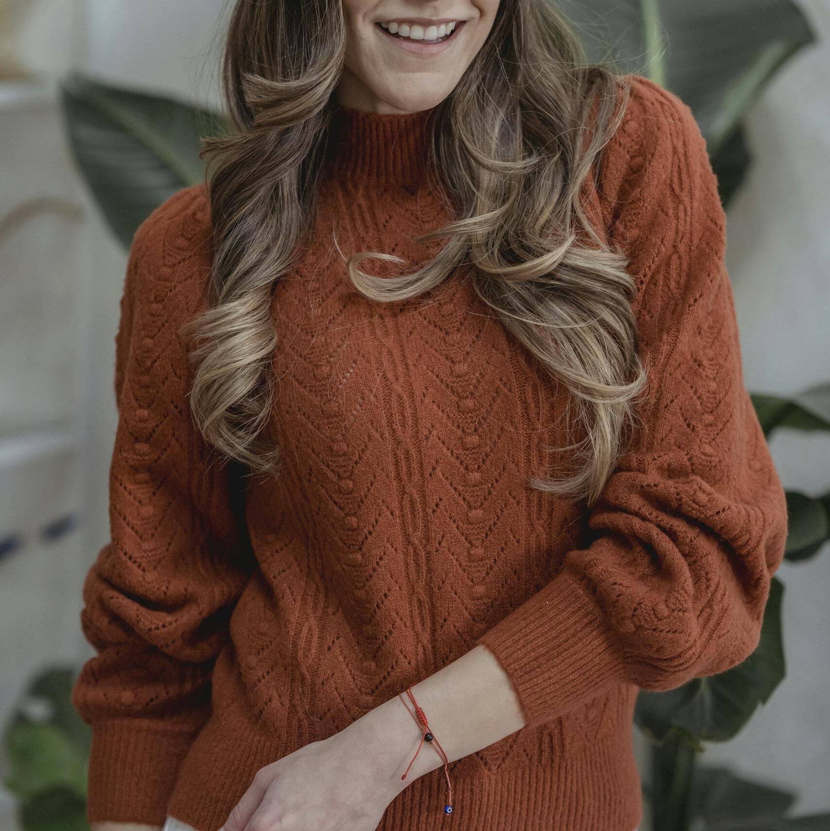Young woman with wavy hair wearing a burnt orange sweater, smiling indoors with green plants.