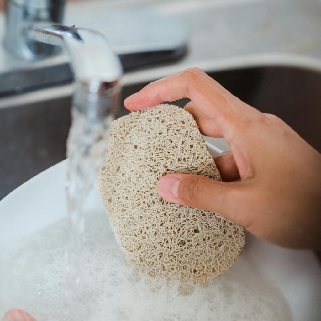Hands washing a plate with a natural loofah sponge under running water in a kitchen sink.