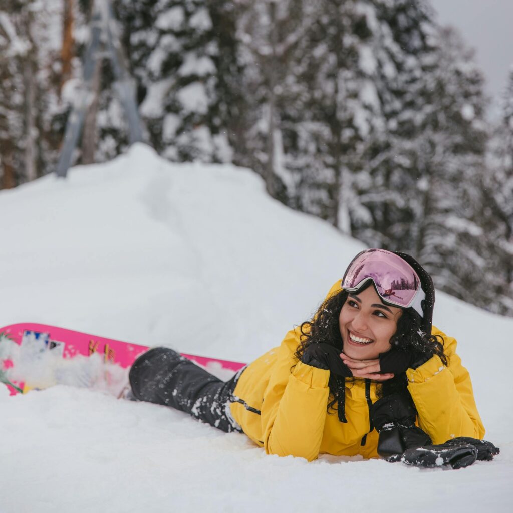 Smiling woman in a yellow jacket lying on snow with snowboard, enjoying winter outdoors.