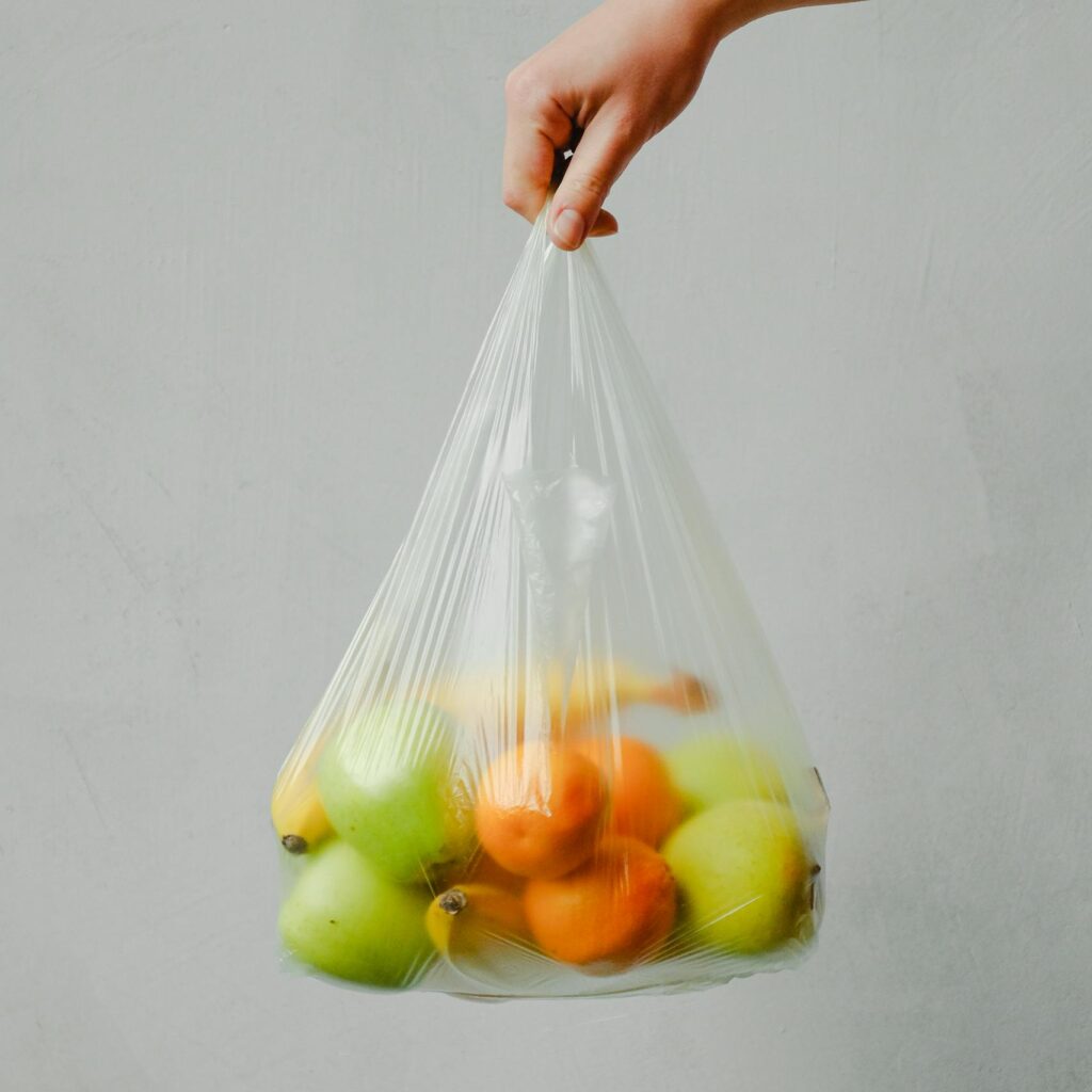 A hand holding a transparent plastic bag full of fresh citrus fruits against a neutral background.