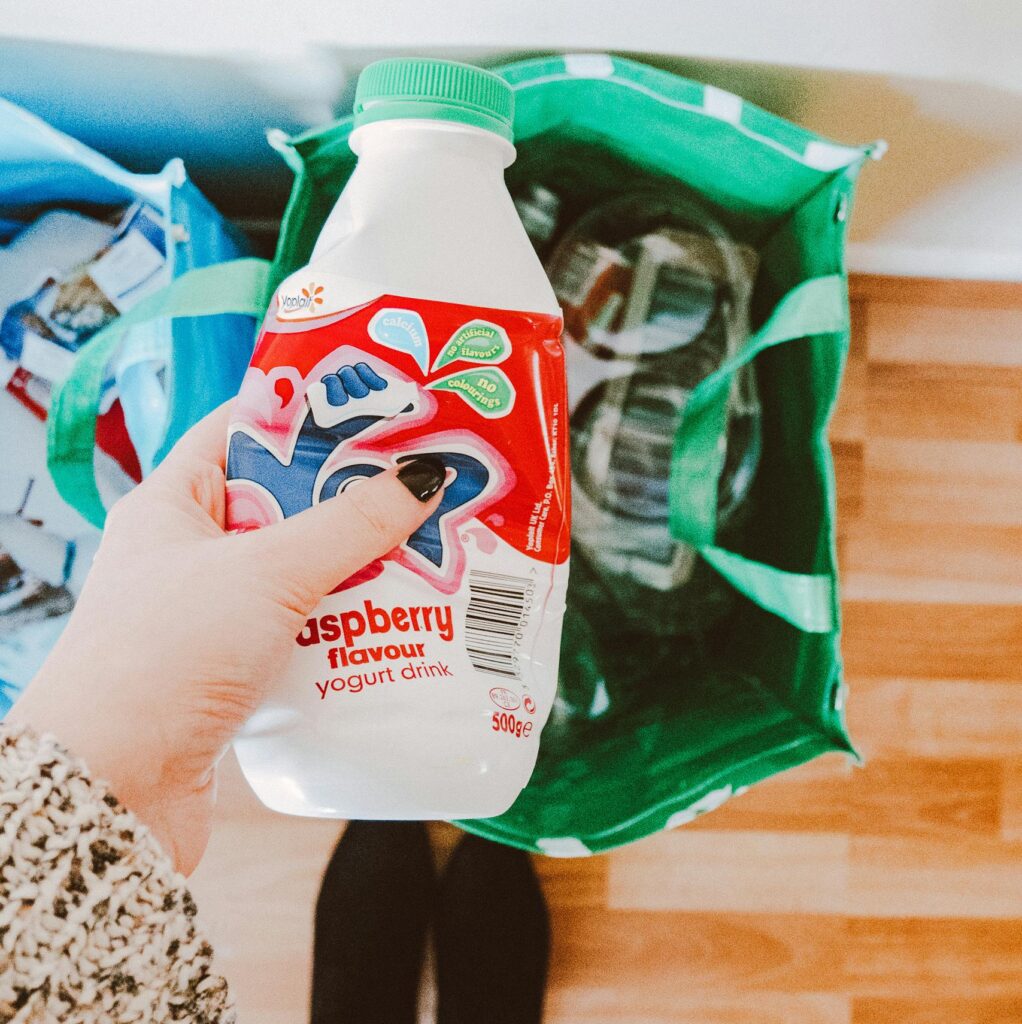 A person recycling a raspberry yogurt drink bottle into a green recycling bag.
