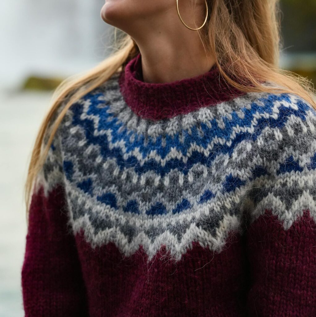 Woman in Icelandic wool sweater smiling by a scenic waterfall in Akureyri, Iceland.