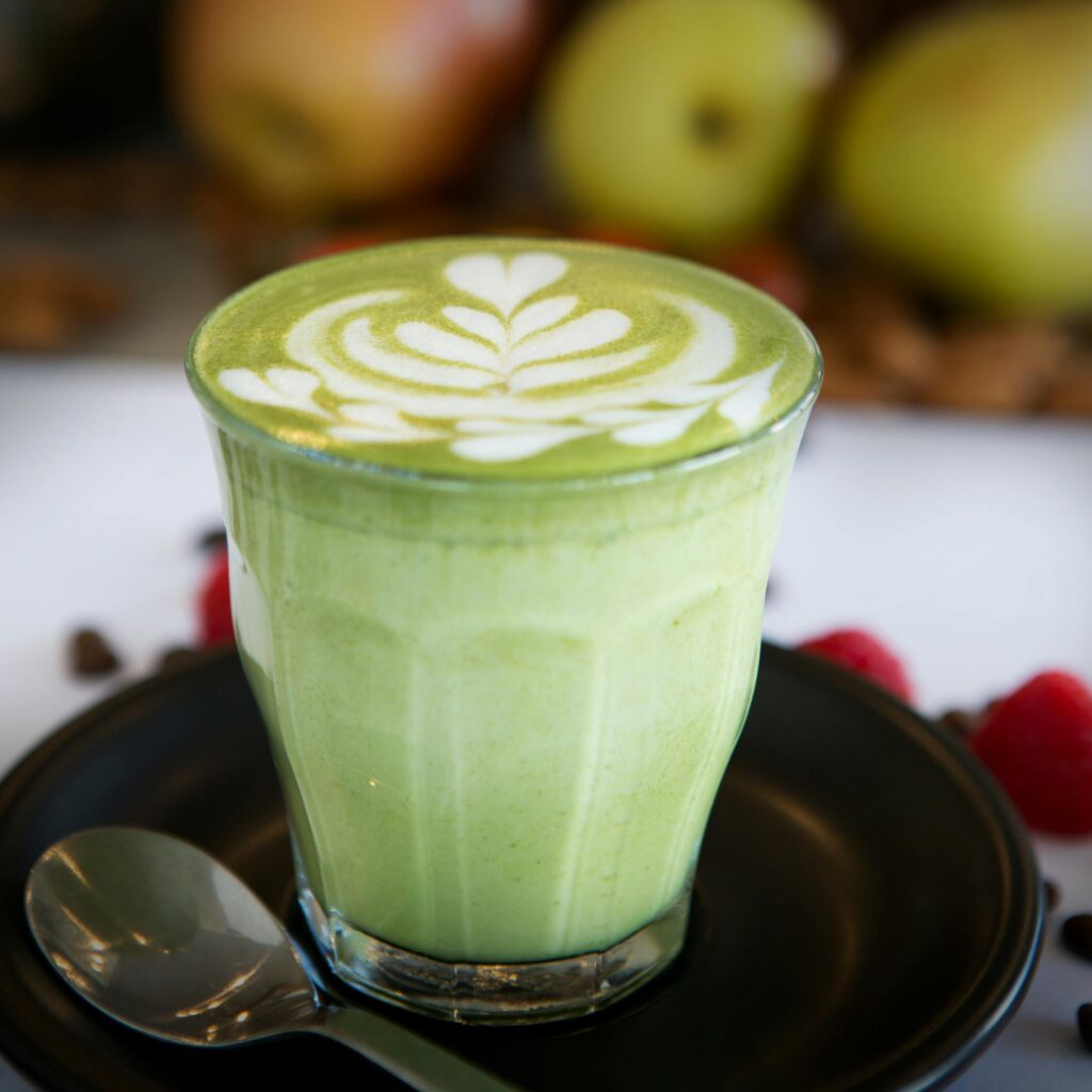 A beautifully crafted matcha latte with latte art in a glass, surrounded by fresh apples and raspberries, perfect for coffee lovers.