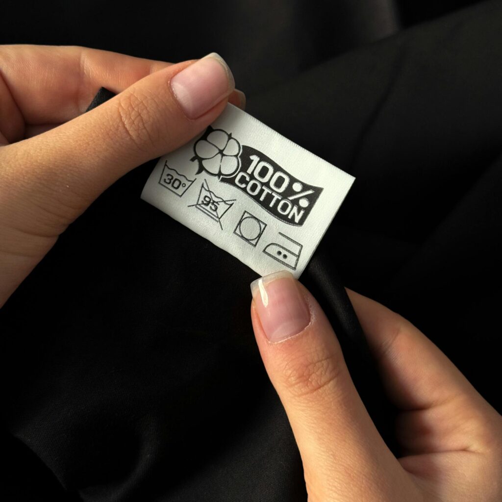 Hands of a woman examining a 100% cotton tag on clothing against a dark background.