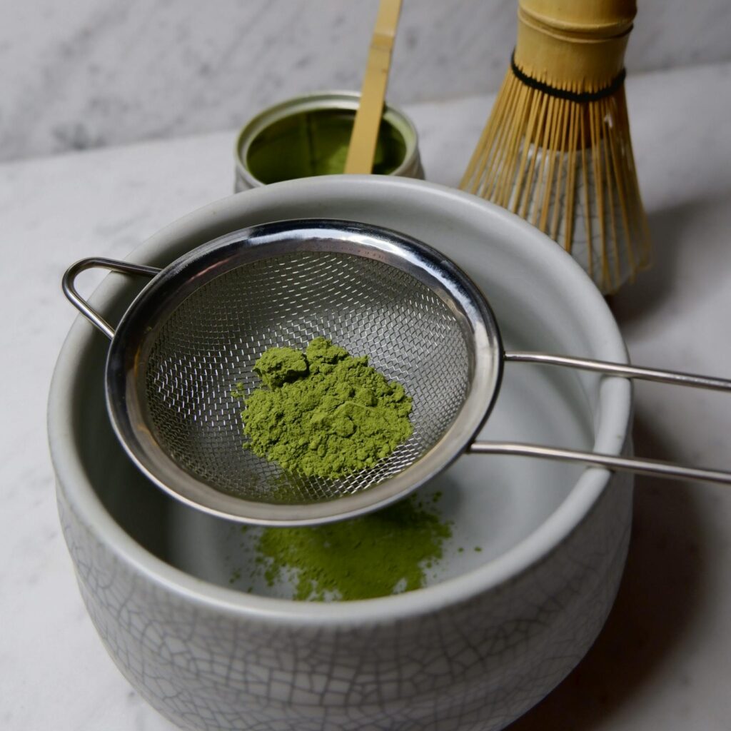 Matcha tea being sifted with a strainer over a bowl, featuring a bamboo whisk and scoop on a marble table.