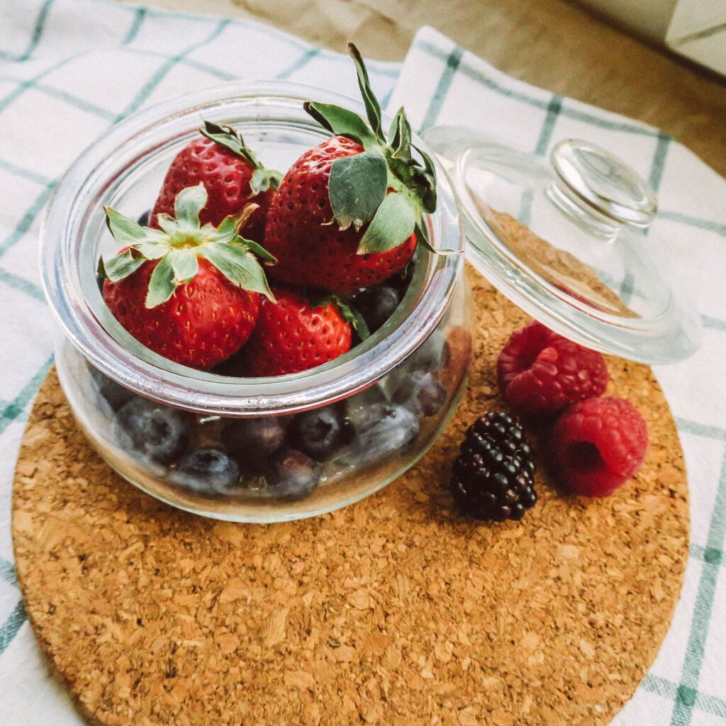 A close-up of fresh strawberries, blueberries, and raspberries in a glass jar on a cork mat.
