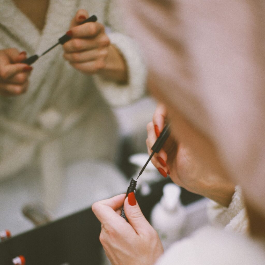 A woman applies makeup in front of a bathroom mirror, focusing on her eyes and reflection.