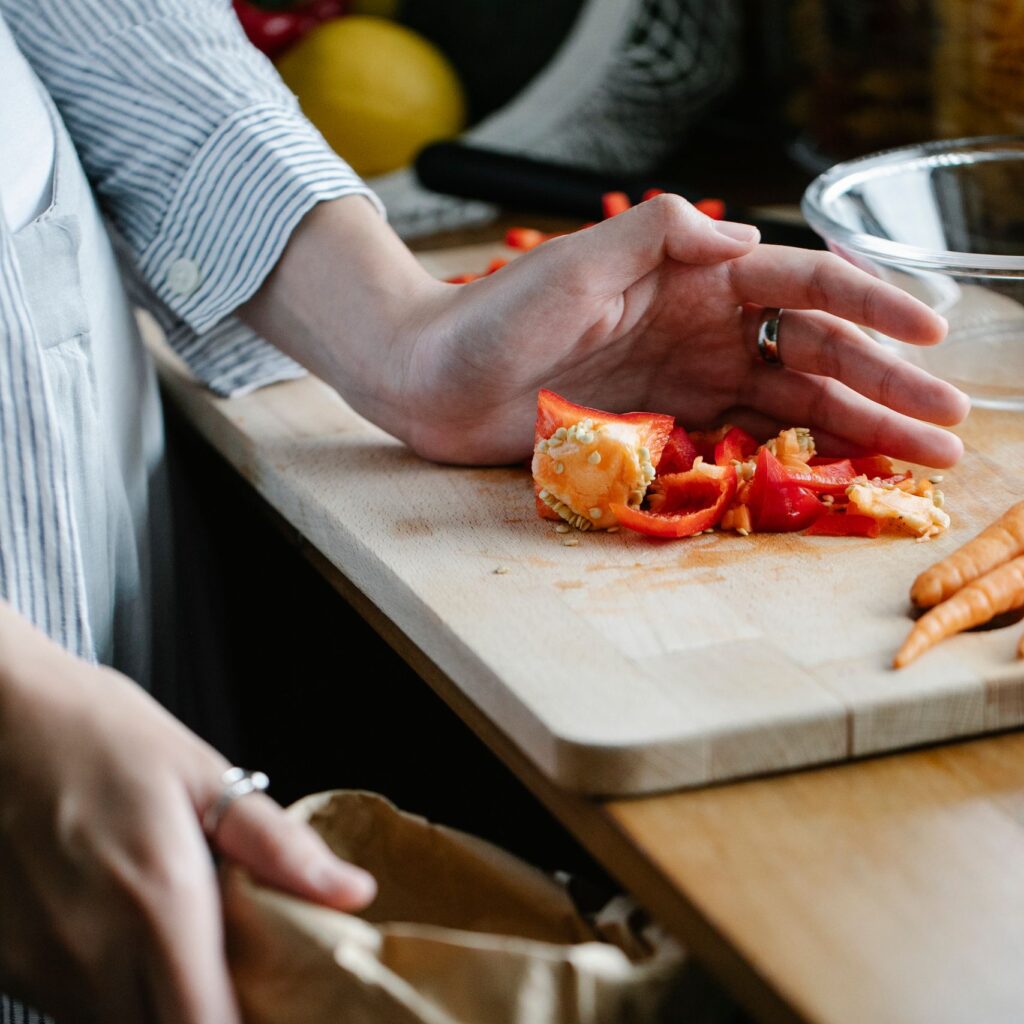 Crop anonymous housewife throwing vegetable leftovers on chopping board while cooking in light kitchen