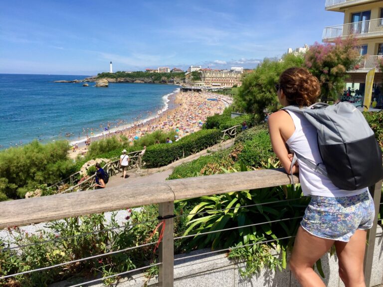 A young woman with a backpack gazes over the bustling beach of Biarritz, France on a sunny day.