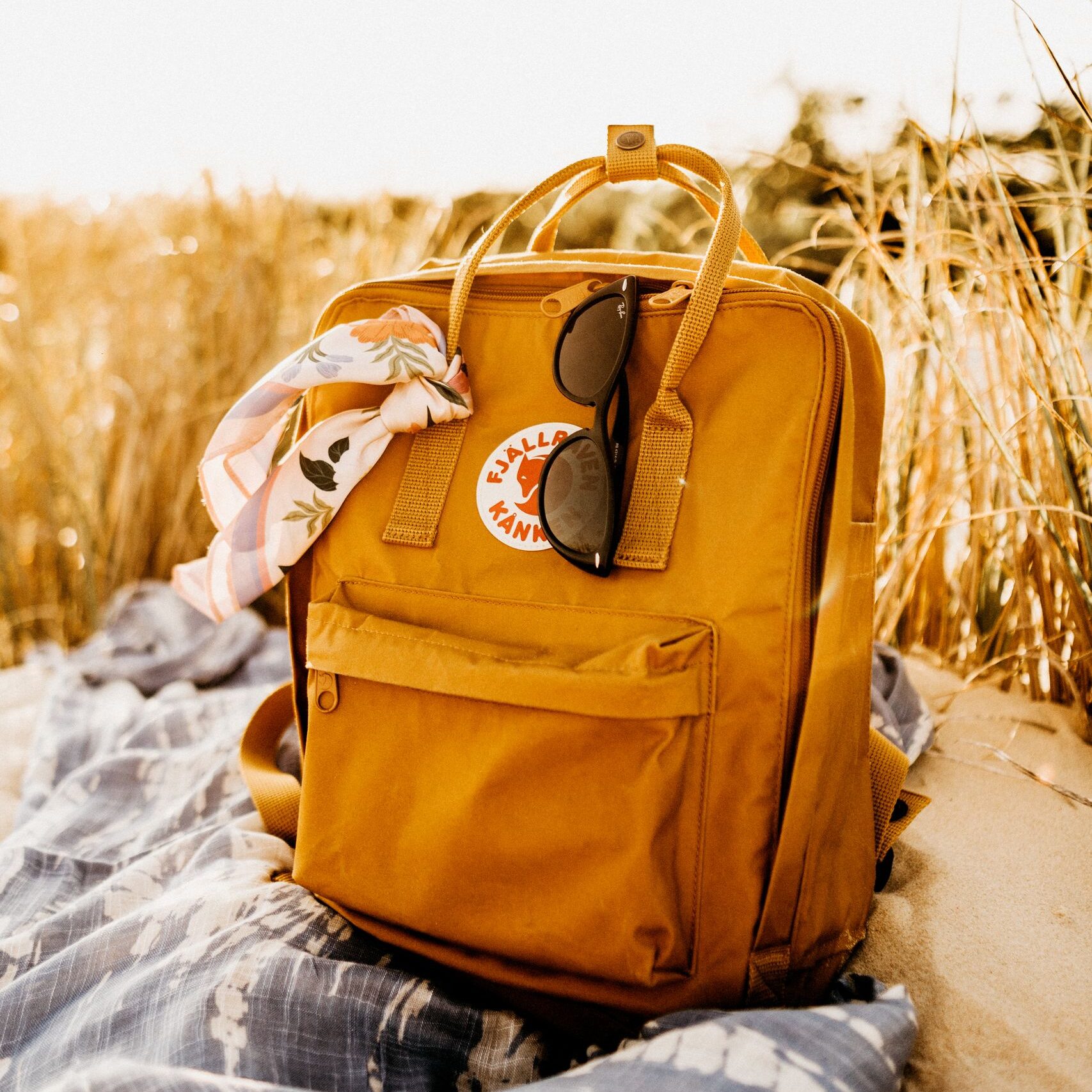 Composition of yellow backpack with sunglasses and handkerchief placed on blanket on sand beach against blurred grass and sky background