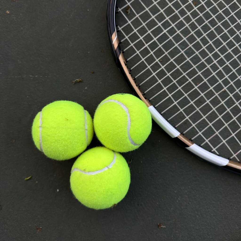 Tennis equipment including three bright tennis balls and a racket on the court surface.