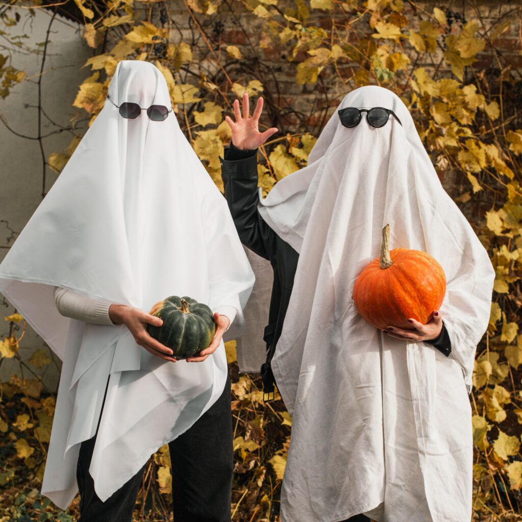 Two people in ghost costumes holding pumpkins amidst fall leaves outdoors.